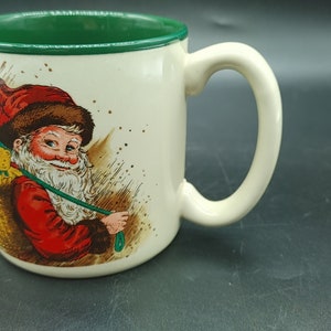 Op de afbeelding: Witte keramische mok met een groene rand. De mok heeft een feestelijke illustratie van Sinterklaas die een zak met speelgoed draagt.