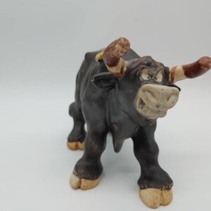 Peut inclure: Une figurine de taureau en céramique noire avec des cornes brunes et blanches. Le taureau a une expression de dessin animé avec une grande bouche et de grands yeux.