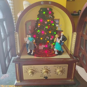 May include: A Christmas-themed musical display with a retro radio design. The display features a decorated Christmas tree, figurines of people dancing, and a control panel labeled "Retro Songs". The display has a wooden cabinet with opening doors.
