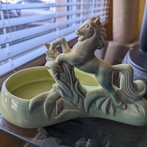 Pu&ograve; includere: Un vaso da fiori in ceramica verde a forma di cavallo con un design a foglie. Il vaso ha una ciotola poco profonda per contenere le piante.