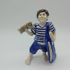 Peut inclure: Une figurine en céramique d'un garçon portant une chemise rayée bleu et blanc et un short bleu. Il tient un journal roulé dans une main et un sac dans l'autre. La figurine a un visage fantaisiste et expressif.