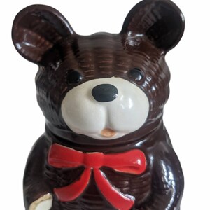 May include: A brown ceramic teddy bear cookie jar with a red bow. The bear has a white face with black eyes and a black nose.