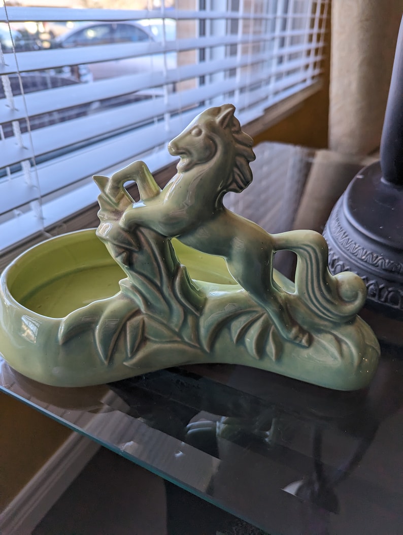 Pu&ograve; includere: Un vaso da fiori in ceramica verde a forma di cavallo con un design a foglie. Il vaso ha una ciotola poco profonda per contenere le piante.