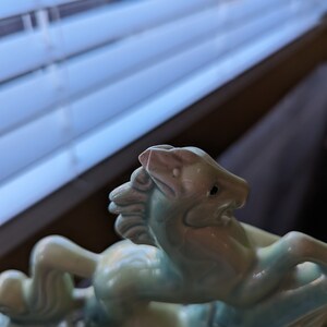 Pu&ograve; includere: Una statuetta in ceramica di un cavallo blu chiaro con criniera e coda bianche. Il cavallo si alza sulle zampe posteriori e ha il naso rosa.