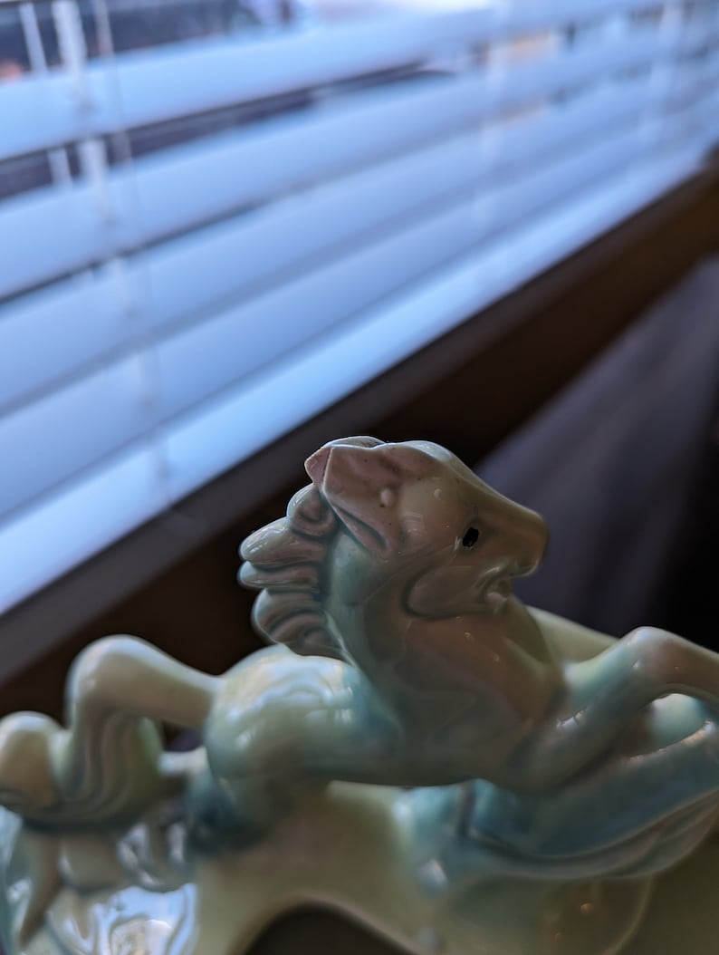 Pu&ograve; includere: Una statuetta in ceramica di un cavallo blu chiaro con criniera e coda bianche. Il cavallo si alza sulle zampe posteriori e ha il naso rosa.