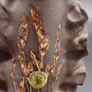 Puede incluir: Un jarrón de cerámica marrón con una superficie texturizada y un diseño en relieve de espigas de trigo. El jarrón tiene una pegatina dorada con el texto "Töpfer Julchen" y "Handarbeit".