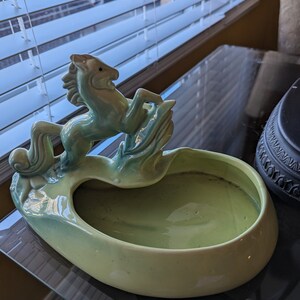Pu&ograve; includere: Un vaso da fiori in ceramica verde a forma di ciotola con una statuetta di cavallo in cima. Il cavallo si alza sulle zampe posteriori e ha una criniera e una coda fluenti.