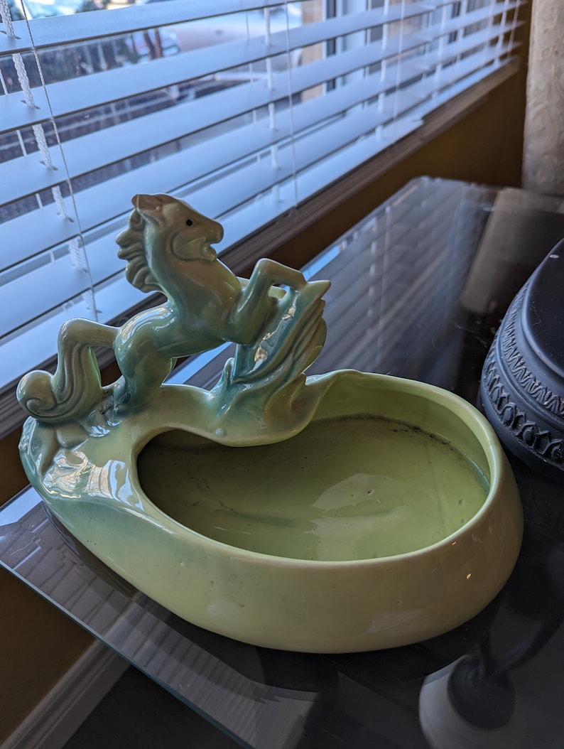 Pu&ograve; includere: Un vaso da fiori in ceramica verde a forma di ciotola con una statuetta di cavallo in cima. Il cavallo si alza sulle zampe posteriori e ha una criniera e una coda fluenti.