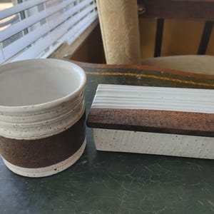 Peut inclure: Un récipient en céramique et une boîte rectangulaire avec un couvercle en bois. Le récipient est blanc avec une bande marron et des détails mouchetés. La boîte est blanche avec une surface texturée et un couvercle en bois brun foncé.