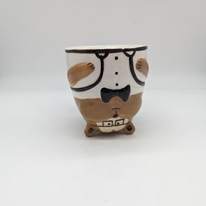 Könnte beinhalten: Eine Keramiktasse in Form eines braunen Bären, der einen weißen Smoking mit schwarzer Fliege trägt. Der Bär steht auf dem Kopf, die Pfoten in der Luft.