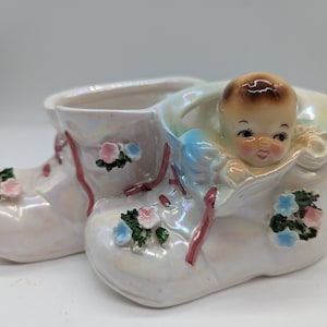 May include: A white ceramic baby shoe planter with a baby's face peeking out of the top. The shoe is decorated with pink and blue flowers.