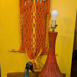 May include: A red ceramic table lamp with a geometric pattern, a white light bulb, and a wooden base. The lamp is next to a teal blue ceramic duck figurine and a macrame wall hanging with orange and yellow yarn.