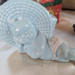 Peut inclure: Une figurine en céramique bleu clair d'un enfant portant un grand chapeau à larges bords. La figurine est couchée sur le côté, la tête reposant sur son bras. Le visage de l'enfant est visible et il a une expression douce.