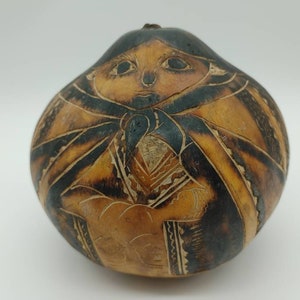Puede incluir: Una calabaza tallada a mano con un diseño marrón oscuro y negro que representa a una persona con un chal. La calabaza es de color marrón claro y tiene una forma redonda y lisa.