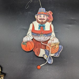 May include: A wooden Thanksgiving-themed pull toy featuring a man in a pilgrim hat holding a pumpkin and a basket of corn. The man has a red beard and a blue bow tie. The toy is painted in bright colours and has a red pull string.
