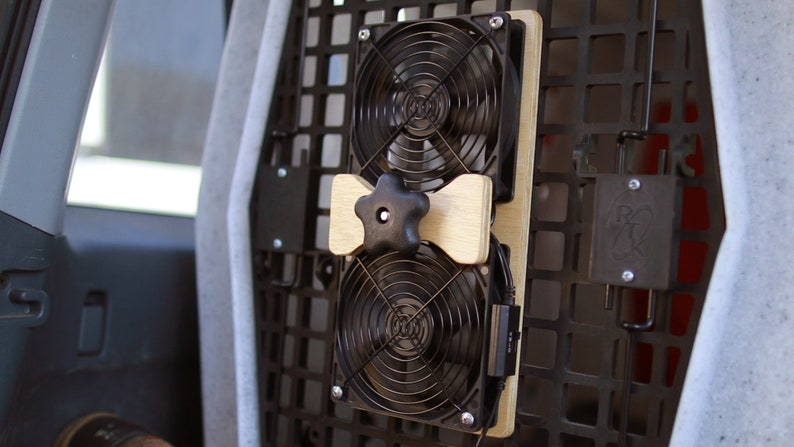 May include: Two black computer fans mounted on a wooden board, secured with a black star-shaped knob. The fans are attached to a black metal grid, likely for vehicle use. A power cord is visible.