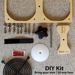 May include: DIY kit for 120 mm fans. Includes a light wood frame with two circular openings, screws, a black fan guard, a white disc, a black knob, and a velcro strap. The text "AirFlow Dually" and "OakLines" are visible.