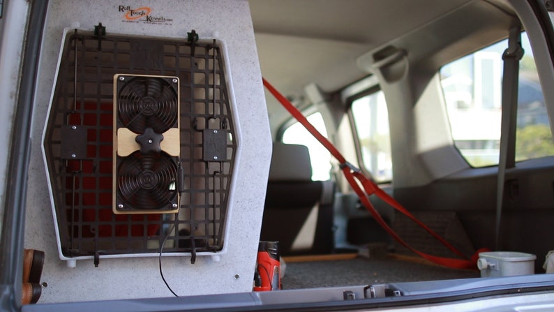 May include: A dog crate with a built-in fan system, installed in the back of a vehicle. The crate is white with a black wire mesh front. The fan system has two black fans and a wooden mounting plate. Red straps secure the crate.