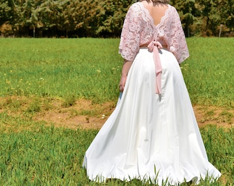 Op de afbeelding: Een vrouw draagt een roze kanten top met een witte rok en een roze lint geknoopt in de taille. Ze staat in een veld met groen gras.