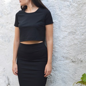 Op de afbeelding: Een zwarte crop top en bijpassende potloodrok. De outfit is gemaakt van een effen, donkere stof. De persoon draagt grote zilveren oorringen. De achtergrond is een witte muur met textuur en groene bladeren.