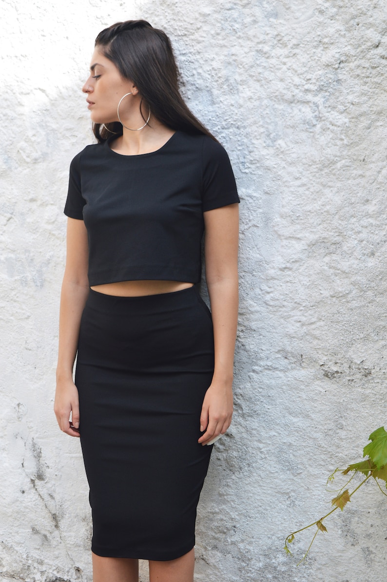 Op de afbeelding: Een zwarte crop top en bijpassende potloodrok. De outfit is gemaakt van een effen, donkere stof. De persoon draagt grote zilveren oorringen. De achtergrond is een witte muur met textuur en groene bladeren.