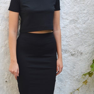 Op de afbeelding: Een zwarte tweedelige outfit bestaande uit een crop top met korte mouwen en een knielange potloodrok. De bijpassende set is gemaakt van een effen, donkere stof. Het model draagt grote oorringen.