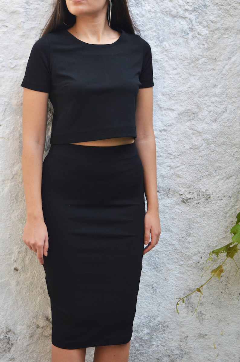 Op de afbeelding: Een zwarte tweedelige outfit bestaande uit een crop top met korte mouwen en een knielange potloodrok. De bijpassende set is gemaakt van een effen, donkere stof. Het model draagt grote oorringen.
