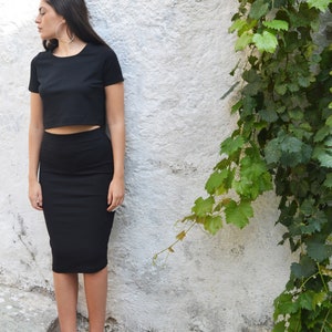 Op de afbeelding: Een zwarte crop top en potloodrok set met rode hakken. De outfit wordt gedragen met grote zilveren oorringen. Het model staat voor een witte muur met groene wijnranken.