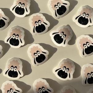 May include: A collection of 15 pink and white sheep stickers with black eyes and noses. The stickers are arranged in a pattern on a white background.