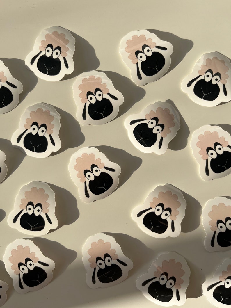 May include: A collection of 15 pink and white sheep stickers with black eyes and noses. The stickers are arranged in a pattern on a white background.
