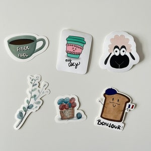 May include: A set of six stickers featuring various designs, including a coffee cup with the text "my cozy", a sheep with big eyes, a cup of coffee with the text "fiber fuel", a sprig of eucalyptus leaves, a basket of yarn, and a slice of bread with a beret and the text "bonjour!"