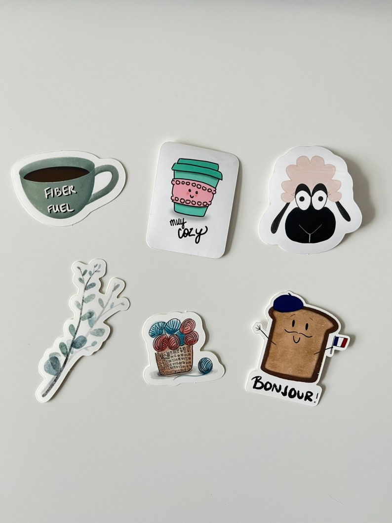 May include: A set of six stickers featuring various designs, including a coffee cup with the text "my cozy", a sheep with big eyes, a cup of coffee with the text "fiber fuel", a sprig of eucalyptus leaves, a basket of yarn, and a slice of bread with a beret and the text "bonjour!"