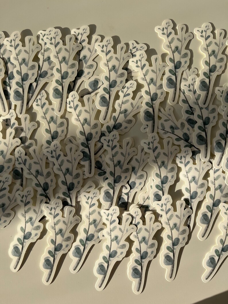 May include: A collection of gray and white watercolor stickers featuring a simple leafy design. The stickers are arranged in a random pattern on a white surface.
