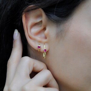 May include: A pair of gold hoop earrings with small pink heart-shaped stones. The earrings are shown in a person's ear.