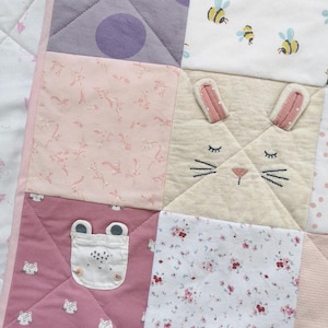 May include: A pastel patchwork quilt with squares of fabric in pinks, purples, and cream. The quilt design includes a bunny face, bees, floral patterns, and polka dots. The quilt is likely for a baby or child, with a soft and inviting aesthetic.