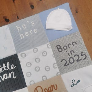 May include: A patchwork baby blanket featuring various textured squares in shades of blue, grey, white, and tan. The blanket includes embroidered text reading he's here, Born in 2025, and Leo, alongside patterns of giraffes and lions, with a white baby hat.