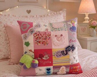 May include: A square patchwork throw pillow featuring various pink, white, and yellow fabric squares with embroidered designs, including a butterfly, a fairy, Piglet, and a small plush green and purple animal attached to the front surface.