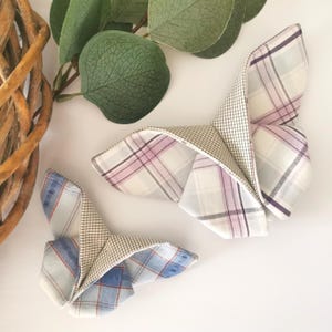 May include: Two decorative butterflies crafted from folded plaid fabric. One butterfly displays a white, pink, and purple plaid pattern, the other a blue and red plaid design. The butterflies are set against a white background with green leaves.
