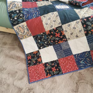 May include: A patchwork quilt featuring squares in red, blue, white, and black, with floral and geometric patterns. The quilt has a blue border and is draped over a bed. The quilt's dimensions are approximately 150 cm x 200 cm.