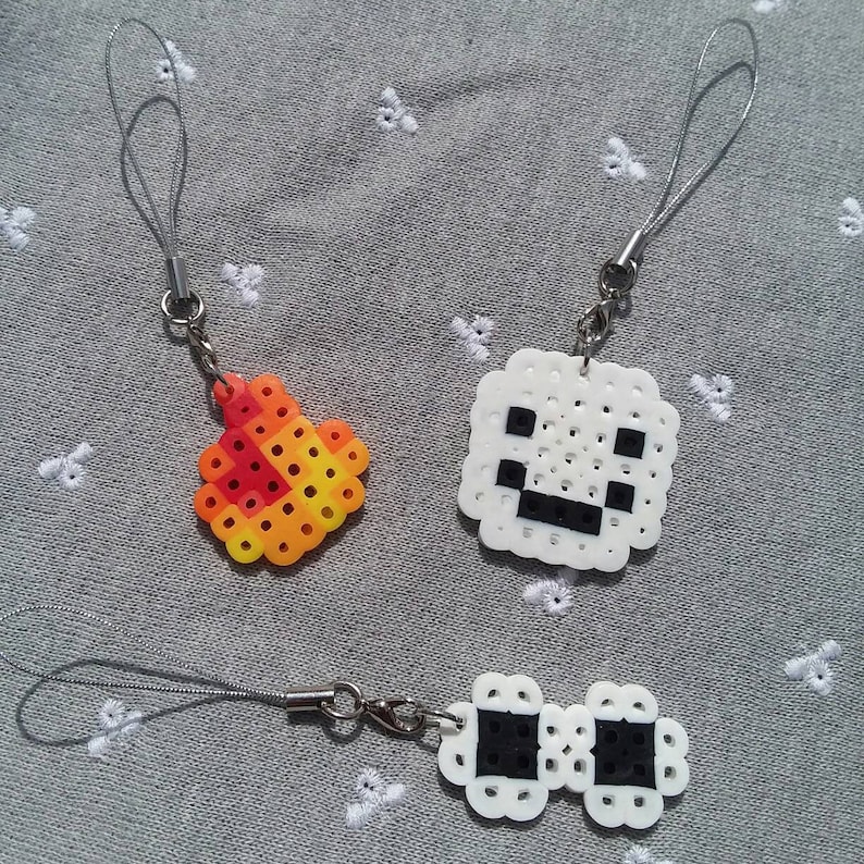 May include: Three white pixel art keychains with black details. One keychain is a square with a face, another is a flame, and the last is a pair of glasses.