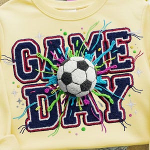 Könnte beinhalten: Hellgelbes Sweatshirt mit dem gestickten Schriftzug "GAME DAY" in Marineblau mit roter Umrandung. Ein Fußball befindet sich in der Mitte des Designs, umgeben von farbenfrohen Fadenakzenten. Das Sweatshirt hat lange Ärmel.