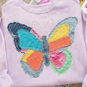 Könnte beinhalten: Ein helllila Sweatshirt mit einem großen, bunten Schmetterling-Applikation. Der Schmetterling besteht aus Stoffflicken in Blau, Gelb, Orange, Rosa und Grün. Das Sweatshirt hat lange Ärmel und einen Rundhalsausschnitt.