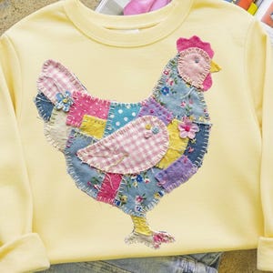Peut inclure: Un sweat-shirt jaune clair orné d'un motif de poule en patchwork. La poule est composée de divers motifs de tissus, dont des motifs floraux, à pois et vichy. La poule a une crête rose et un bec jaune. Le sweat-shirt a des manches longues et un col rond.