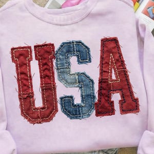 Pode incluir: Um moletom lilás claro com a palavra "USA" costurada na frente. As letras são feitas de tecido vermelho e azul semelhante ao jeans com costuras brancas.