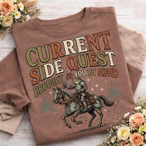 May include: A brown t-shirt with the text "CURRENT SIDE QUEST BROUGHT TO YOU BY ADHD." The graphic features a frog knight on horseback, with castle and star illustrations. The shirt is folded.