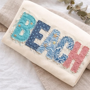 Puede incluir: Un cojín decorativo color crema con la palabra "BEACH" aplicada en varias telas estampadas azules y rosas. Las letras están adornadas con conchas marinas, estrellas de mar y cuentas de perlas, evocando un tema costero.