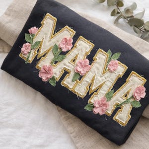 May include: A dark navy blue sweatshirt featuring the word "MAMA" in cream-colored fabric with gold stitching. Pink fabric flowers and green leaves are sewn around the letters.
