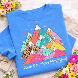 Könnte beinhalten: Blaues Sweatshirt mit farbenfrohem Bergmotiv. Die Grafik zeigt schneebedeckte Gipfel, Herzen und Sterne, mit dem Text "Faith Can Move Mountains" darunter. Das Design umfasst Leopardenmuster und verschiedene Farben.