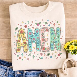 May include: Cream-coloured sweatshirt featuring the word "AMEN" in vibrant floral lettering. The letters are adorned with small dots and surrounded by crosses, hearts, stars, and floral details. The sweatshirt is folded on a wooden surface.