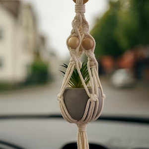 Könnte beinhalten: Ein cremefarbener Makramee-Pflanzenaufhänger mit einem kleinen, grauen Topf, der eine grüne Pflanze enthält. Der Aufhänger hat Holzperlen und einen Holzring an der Oberseite. Die Pflanze hängt vor einem verschwommenen Hintergrund.
